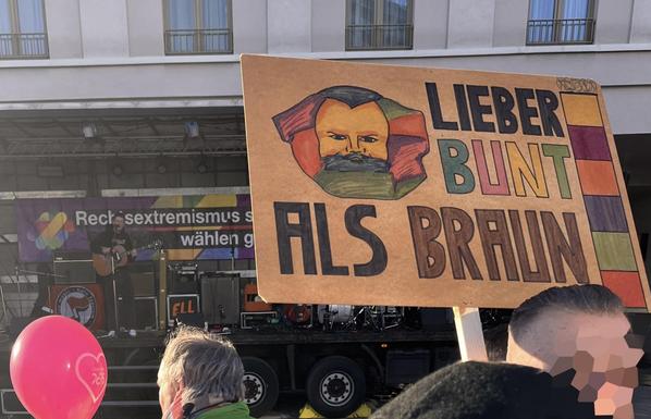 A protest scene featuring a sign with the message "Lieber bunt als braun" ("Better colorful than brown"). The sign includes a colorful illustration of Karl Marx. In the background, there’s a performer with a guitar on stage and a