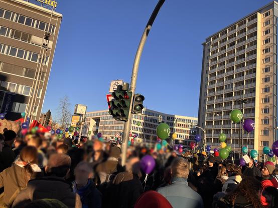 A crowd of people is gathered in a city square, holding colorful balloons. Surrounding buildings are visible, along with traffic lights and a clock. The sky is clear and blue, indicating a sunny day.
