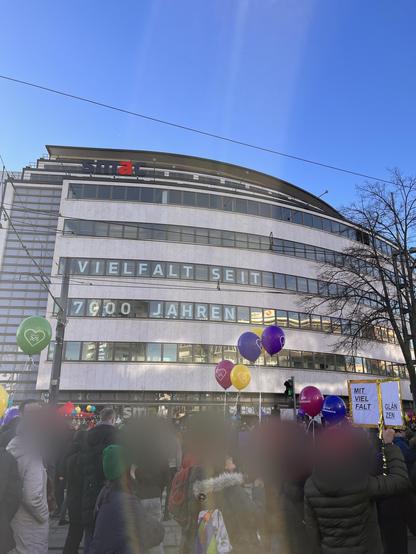 A crowd gathers in front of a building with a clear blue sky above. The building displays the text "Vielfalt seit 7.000 Jahren" on its facade, emphasizing diversity. People hold colorful balloons and signs reading "Mit Vielfalt glänzen."
