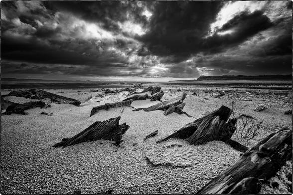 Das Bild zeigt eine schwarz-weiße Landschaftsaufnahme. Im Vordergrund sind Reste von Schiffsplanken auf einem kiesigen oder sandigen Strand verteilt. Der Himmel ist dramatisch mit dichten, dunklen Wolken bedeckt, die eine stürmische Atmosphäre erzeugen. 