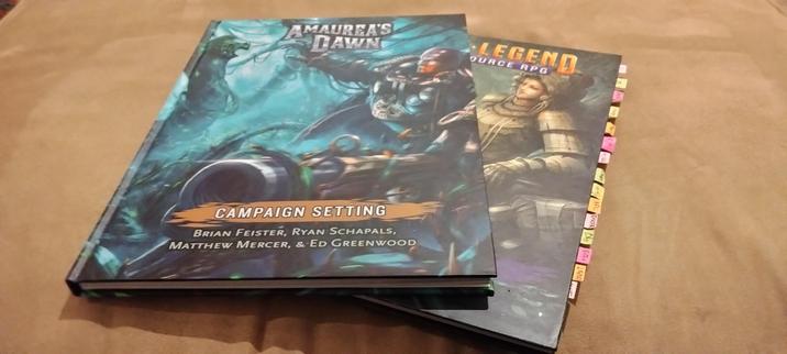 Two books lying askew. On top, "Amaurea's Dawn" campaign setting source book. Below, "Open Legend RPG" core rule book, with multi-color tags emerge from the right side.