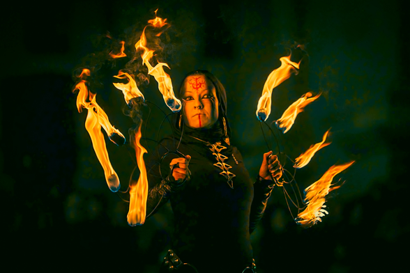 Fotografía de mujer vestida de negro, con joyería plateada y la cara pintada de blanco y negro con rayas y símbolos en rojo. Está sujetando abanicos de manipulación, cada uno de ellos tiene 5 llamas de fuego. Es una fotografía tomada durante un espectáculo de fuego en un mercado medieval.