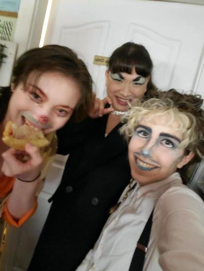 A group photo of Austin Tacious, Kaleidescope Crush, and Spectra Savant before the show. Austin is dressed as the Snowmiser. Kaleidoscope is dressed as the Heatmiser, in a bright orange shirt, with red eyebrows, a green moustache, and a sparkly red nose. Spectra has blue eyeshadow, arched eyebrows, and smooth bangs. She's wearing a diamond necklace and a black coat. It's also worth noting that Kaleidoscope is in the middle of eating a slice of pizza.