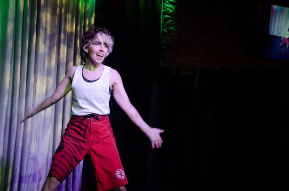 Austin Tacious, onstage at the Junction. He's stepping animatedly, dressed as Rocky Balboa in red sports shorts with black stripes, a wifebeater, and tan bandages around his hands. His eyeshadow is tiger-striped, and his contouring is an orange sort of colour.