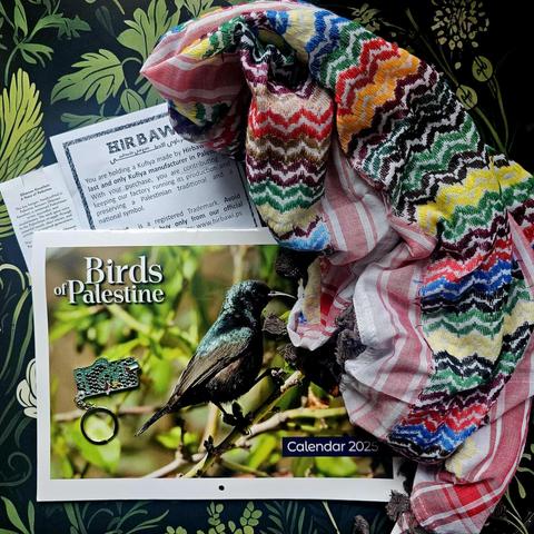 Photo of a keffiyeh in the colora of the rainbow on the right, a tekstsheet with 'HIRBAWI' writen on top of it in atyled letters. In the center if the picture a calander is displayed with the tekst 'Birds of Palestine - calendar 2025' woth thw picture of a bird on the cover with on top of that a green colored keychain with the Palestinian flag on it. On the background a green botanical display of illustrated plants and flowers.