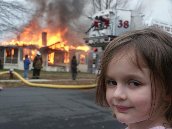 A young girl stands in the foreground with a slight smile as a house burns in the background, engulfed in flames and smoke. Firefighters are seen working nearby, attempting to control the fire.