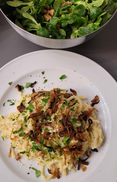 Farbfoto hochkant: Unten im Bild ist ein weißer Teller mit einer Portion Käsespätzle zu sehen, darauf frische Röstzwiebeln, etwas gehackte Petersilie und frisch gemahlener Pfeffer. Oben im Bild ist angeschnitten eine Metallschüssel mit Feldsalat zu sehen, darin ein paar Walnüsse. 