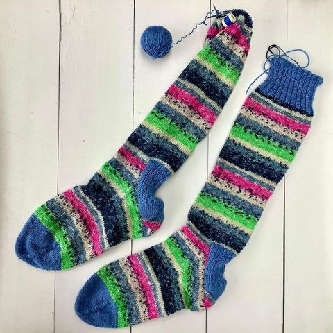 2 socks in green, pink, and white self-striping yarn on a white plank background. One is still missing its ribbing.