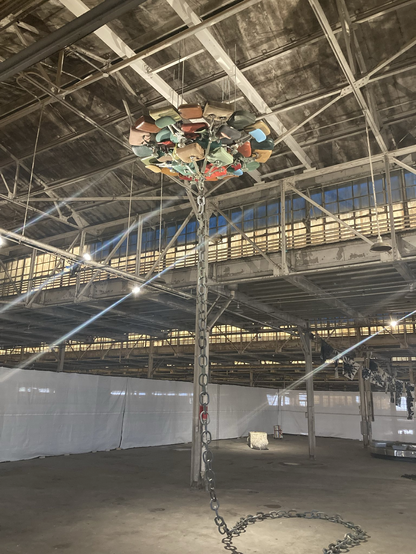Fabrikhalle von innen, Licht kommt durch das Oberlicht im 1. Stock. An der Decke hängt eine große Installation aus Sesseln, von der eine Kette auf den Boden herabhängt.