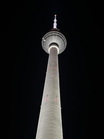 Auf dem Foto sieht man den Berliner Fernsehturm bei Nacht. Der Turm ist von unten aufgenommen, wodurch er imposant und hoch wirkt. Er ist hell beleuchtet, und der dunkle Nachthimmel bildet einen starken Kontrast zur Struktur des Turms. Die roten Markierungen und die Spitze des Turms sind ebenfalls sichtbar.