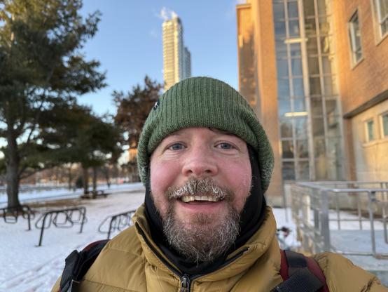 Smiling beared guy in a mustard puff jacket and green toque. Classic selfie shot with buildings and snow in the background. Some ice visible in the beared and bike racks in the distance. 