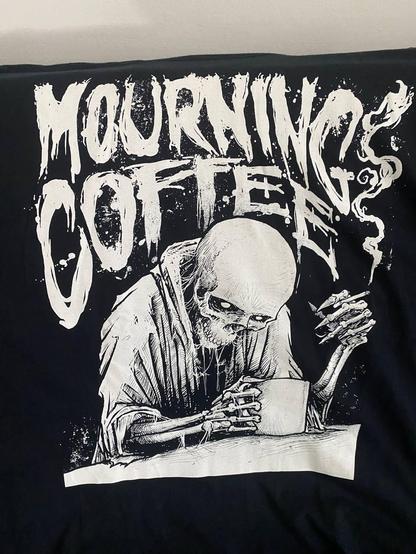 Illustration, weiß auf schwarzem Untergrund. Ein Skelett mit einem Pullover sitzt an einem Tisch und hält in der einen Hand einen Kaffeebecher und in der anderen eine Zigarette.
Dazu ein rauchig verwischter Schriftzug: Mourning Coffee