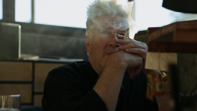 A screenshot from the film DAVID LYNCH: THE ART LIFE showing Lynch in a contemplative moment, holding a cigarette and looking upwards.