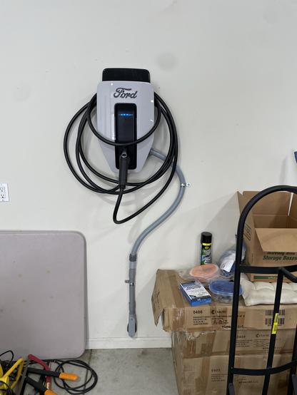 An electric vehicle charging station mounted on a wall, featuring a Ford logo. The station has a coiled power cable and is located in a garage with cardboard boxes and other items scattered around.