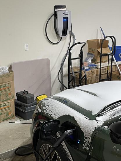 An electric vehicle is charging in a garage, with a wall-mounted Ford charging station nearby. Snow is visible on the vehicle's hood and around the charging port. Various storage boxes and cleaning supplies are visible in the background.