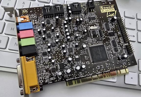 Creative Sound Blaster Live! PCI sound card, model SB0060. The card features multiple capacitors, an EMU10K1 audio processor, gold-plated PCI contacts, and a variety of input/output ports, including 3.5mm jacks and a proprietary yellow connector. The 'Sound Blaster Live!' branding is printed on the PCB. The card is resting on a white keyboard.