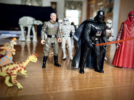 A green Dilophosaurus with brown spots stands alongside: a tan Parasaurolophus, a brown Dilophosaurus, a light tan Pachycephalosaurus, and a very short Brachiosaurus. They stand in a line watching a parade. Darth Vader leads the column, lightsaber in hand with an Imperial Officer to his right and a red Imperial Guard on his left. A large column of white Imperial Stormtroopers follow with a few TIE Fighter pilots behind them. An Imperial AT-AT walker follows at the parade's end.  