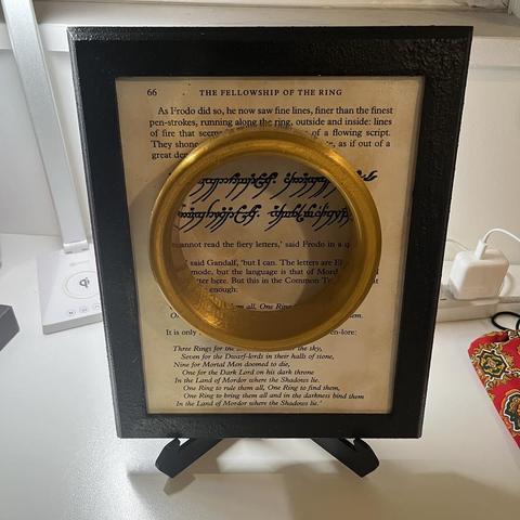 A picture frame with a rocky texture painted black. It contains a page from the Lord of the Rings first book, the part where Gandalf describes the ring to Frodo and quotes the elvish poem.
The ring is glued on top of the page at the center of the frame.