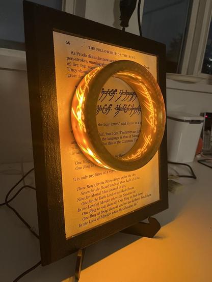 A picture frame with a rocky texture painted black. It contains a page from the Lord of the Rings first book, the part where Gandalf describes the ring to Frodo and quotes the elvish poem.
The ring is glued on top of the page at the center of the frame, but it's lit from the inside so there is some scripting visible around the ring with a yellow/orange hue.