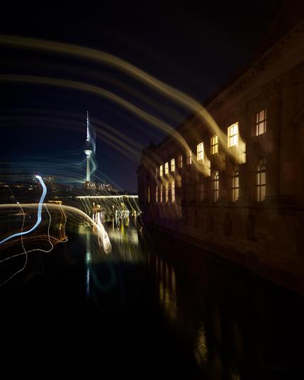 A nighttime scene in Berlin, captured along the Spree River. On the left side of the image, the illuminated Berlin TV Tower is visible, surrounded by flowing, curved light trails created through long exposure. On the right side, the lit windows of a historic building reflect on the calm water of the Spree. The image combines architecture, motion, and light in an artistic composition.