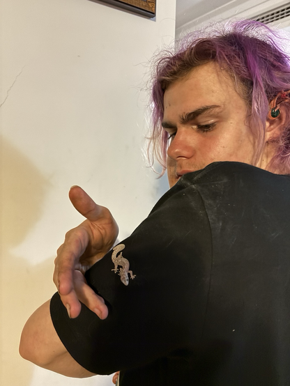 A teenage boy with purple hair, looking over his shoulder at a small pink gecko on his black tshirt sleeve.