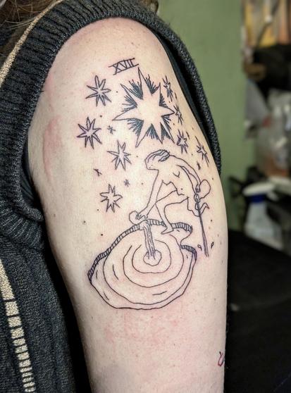 Tattoo on upper arm. The design is based on a tarot card (The Star) and features a naked woman pouring water in a pond and on the ground while sitting on one leg under a big sky of stars. It says XVII on top, a reference to the tarot card. The style is a bit wonky and playful