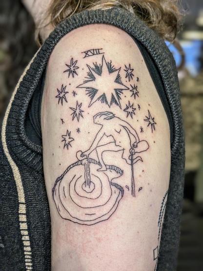 Tattoo on upper arm. The design is based on a tarot card (The Star) and features a naked woman pouring water in a pond and on the ground while sitting on one leg under a big sky of stars. It says XVII on top, a reference to the tarot card. The style is a bit wonky and playful