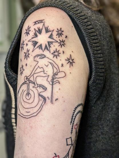 Tattoo on upper arm. The design is based on a tarot card (The Star) and features a naked woman pouring water in a pond and on the ground while sitting on one leg under a big sky of stars. It says XVII on top, a reference to the tarot card. The style is a bit wonky and playful