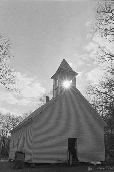 A black-and-white photo of a historic wooden church with the sun shining through its bell tower, surrounded by bare trees.
