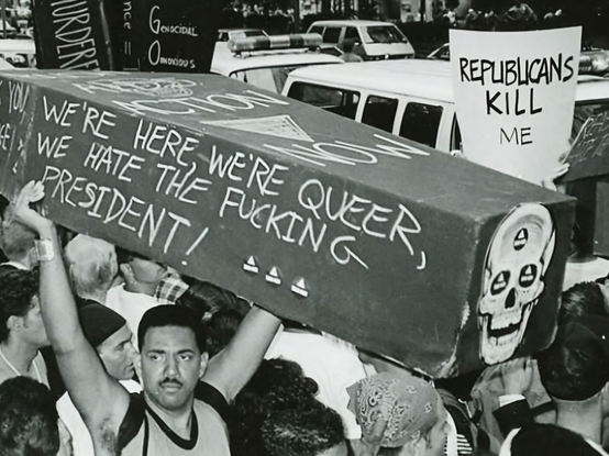 A 1990 photo of an Act Up protest during the AIDS/HIV crisis. A man (the poet  Assotto Saint)  carries a coffin with a skull on the front and writing on the side:  "We're here we're queer, we hate the fucking president"