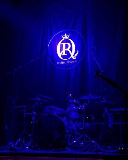 photo of a drum set on an empty stage pre concert. rolling quartz logo on the backdrop.
stage filled with blue lighting.