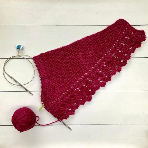 Flat lay view of a knitting project, featuring garter stitch and lace on a circular needle. It's shown on a white plank background.