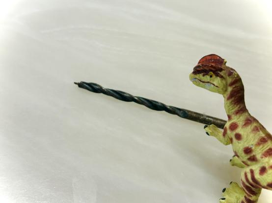 A green Dilophosaurus with brown spots stands before a drill bit on a white, porcelain surface.