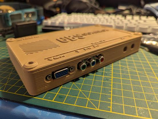 A photograph of a GBS-Control scaler device.  The electronics are installed in a 3D-printed box the color of peanut butter, with legends for the inputs and outputs printed standing out from the surface of the top of the box.  Inputs for VGA and component video are facing the viewer.
