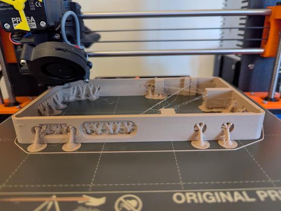 A photograph of the printing bed of a Prusa MK3S+ 3D-printer, most of the way through printing part of the case for the GBS-C upscaler.  The filament material is exceptionally stringy, and fine strings are criss-crossing everywhere.  The print is being done with tree supports, and those combined with the strings are making it look extra-organic.