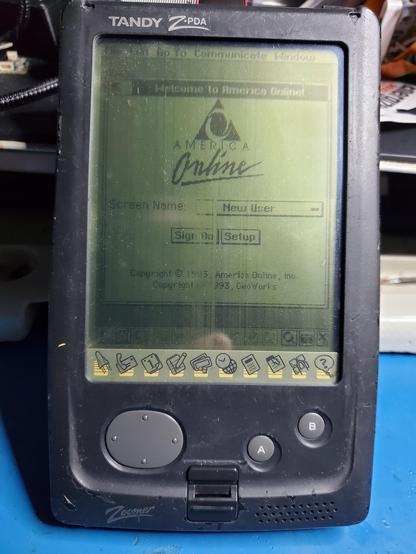Photo of a slate-style PDA with a monochrome LCD display showing the America Online login window. The image on the screen is missing some stripes in an irregular pattern.