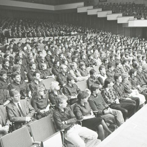 The black and white photo shows a lecture theatre in which many people are sitting and watching an event.