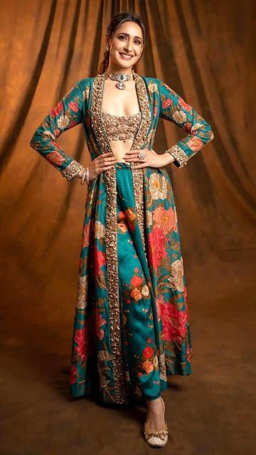 Actress Pragya Jaiswal stuns in a traditional outfit that masterfully combines modern floral patterns with classic design. The ensemble features a sleeveless golden embroidered blouse, a full-length satin skirt with bold red, gold, and cream floral prints, and a regal open-front jacket. Accessorized with a pearl and emerald choker and gold-plated kundan ring, Pragya's look is the epitome of grace and style.