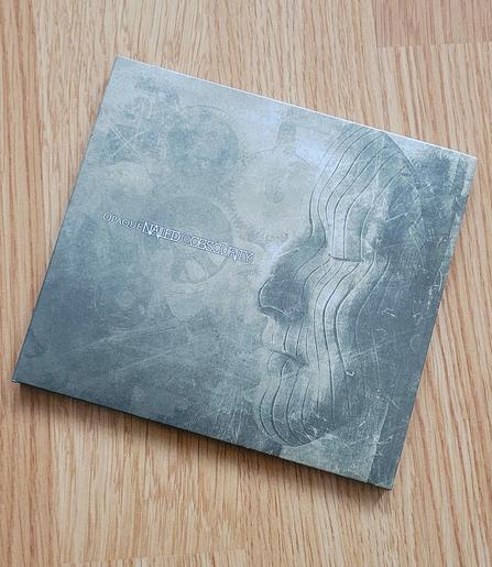A CD album on orange-brown flooring ; album cover is a stone face on the right edge with decorative vertical grooves, looking to the left, where the silhouettes of multiple mechanical gears can be recognised. The whole cover is grey with a greenish tinge. Album title "Opaque" and band name "Nailed to Obscurity" written in the middle with white characters