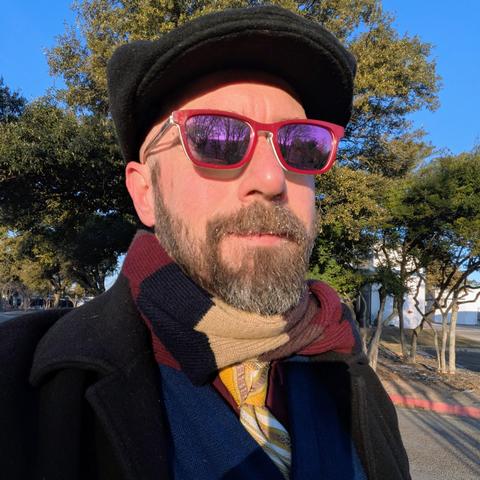 A photo of me, a 46 year old white man with a shaved head, a short cropped brown & grey beard, 

photographed from the chest up in front of some live oak trees in an office park,

wearing

a rectangle glasses with maroon frames, a yellow necktie with white stripes in a trinity knot on a maroon dress shirt under a navy blue cardigan and a black topcoat with a black flat cap and navy blue, tan, and maroon striped scarf