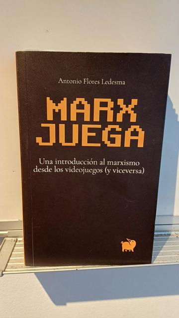 Foto del libro Marx Juega: una introducción al marxismo desde los videojuegos (y viceversa). Apoyado en el estante de la pizarra blanca que tenemos por aquí, en Aula Pública.