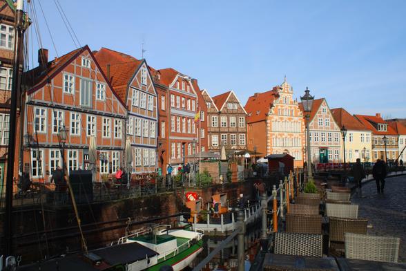 Hafenbecken von Stade mit dahinterliegender Häuserzeile. Die Gebäude haben ganz unterschiedliche Baustile. Auf der rechten Seite stehen entlang des Hafenbeckens ein paar Stühle (Außengastronomie). Zwei Personen gehen die Straße entlang. Die Sonne steht tief und scheint auf die Häuserzeile.
