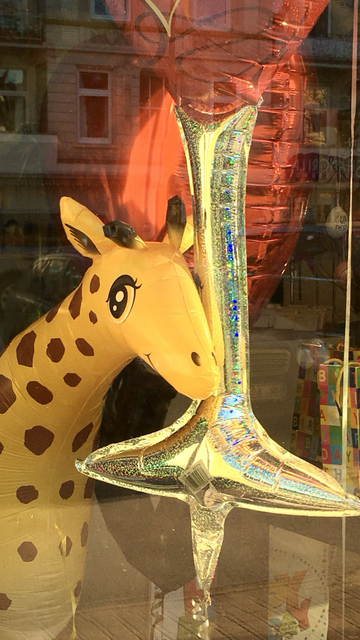 Schaufenster eines Luftballon-Ladens mit einem comic-haften aufgeblasenen Giraffenkopf. Die Giraffe hat große Kulleraugen und lächelt freundlich. Daneben ein undefinierbarer länglicher Folien-Luftballon mit Glitzeroberfläche und dahinter ein großer glänzender Folienluftballon in Rot. - Shop window of a balloon shop with a comic-like inflated giraffe head. The giraffe has big, round eyes and a friendly smile. Next to it is an indefinable, long foil balloon with a glittery surface and behind it a large, shiny red foil balloon.