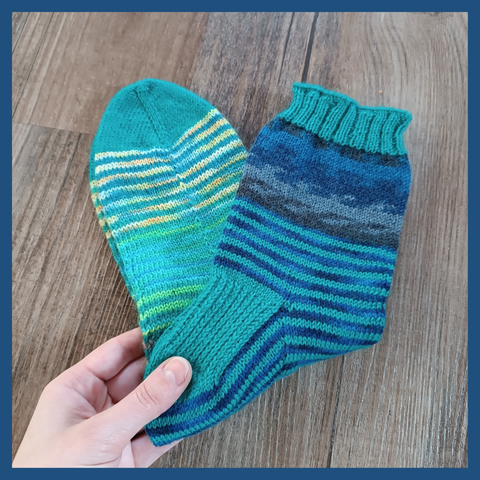 A pair of knitted socks with stripes in different shades of blue, green, yellow and black with a turquoise shade as base stripes. The heel flap, toe and upper ribbing are the same shade of turquoise. A light coloured hand holds the socks into the picture onto a background of a dark wooden floor.