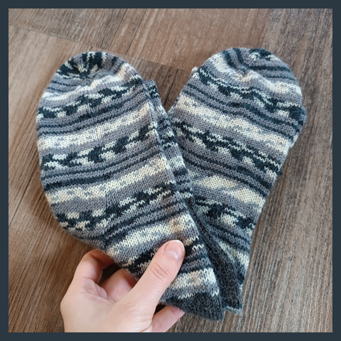 A pair of socks folded into a heart shape, knitted with a self-striping yarn in different shades of grey. A light coloured hand holds them in place on a dark wooden floor.