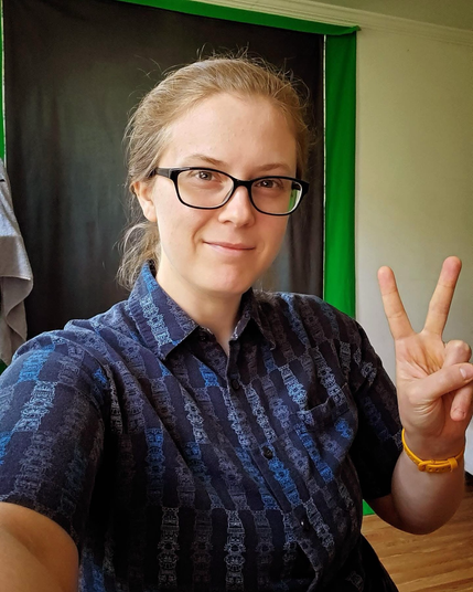 A genderfluid nerd wearing glasses and a dark blue patterned men’s button-up short sleeved shirt from the 90’s, gesturing with a peace sign in front of a greenish blue backdrop with a hint of green screen peeping out on a blank off-white wall