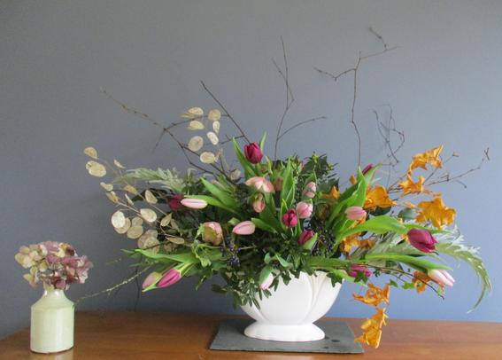 A large flower arrangement in a white vase on a wood table. The arrangement is in an asymmetric fan shape. British grown pink and red tulips are supported by a range of dried and fresh foliage.