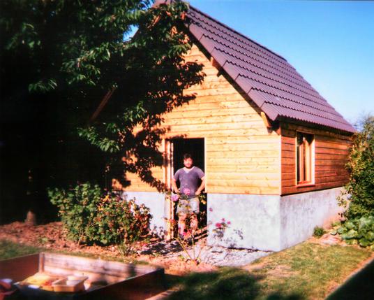 Pascal MOREL, gérant de l'entreprise J.M.B. Construction, posté dans la porte d'une dépendance en forme de petite maison à la base grise sur un demi étage puis en planche en bois verticales sur le reste de la hauteur, au coucher du soleil. Le toit est gris. Devant il y a des parterres de rosiers et un bac à sable et des arbres à gauche.