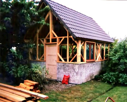 Une dépendance en construction en forme de petite maison à la base grise sur un demi étage puis des poutres en bois sans encore de revêtement sur le reste de la hauteur. Le toit est gris. Devant il y a un tas de planches, à droite un buisson et des arbres à gauche.