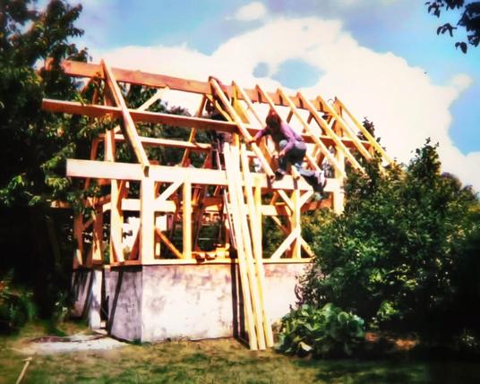 Une dépendance en construction en forme de petite maison à la base grise sur un demi étage puis des poutres en bois sans encore de revêtement et la charpente à nue sur le reste de la hauteur. Dans la charpente Pascal MOREL, gérant de l'entreprise J.M.B. Construction, est perché au milieu. Devant à droite il y a un buisson et des arbres à gauche.